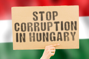 The phrase " Stop Corruption in Hungary " on a banner in men's hand with blurred Hungarian flag on the background. Forbidden. Prevent. Wealth. Offence. Corruptness. Economy. Corruptible. Political