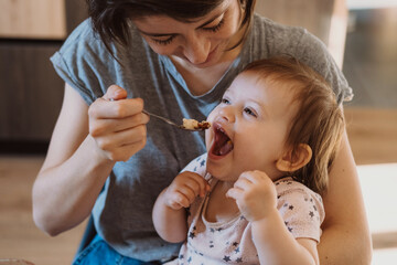 Mother feeding with spoon her little baby at home. Healthy young girl. Baby care. Family care.