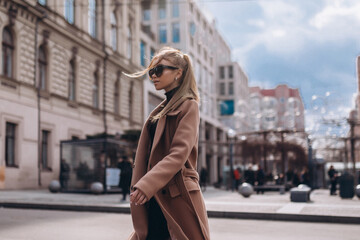 Young beautiful girl in a beige coat and sunglasses walks through the city
