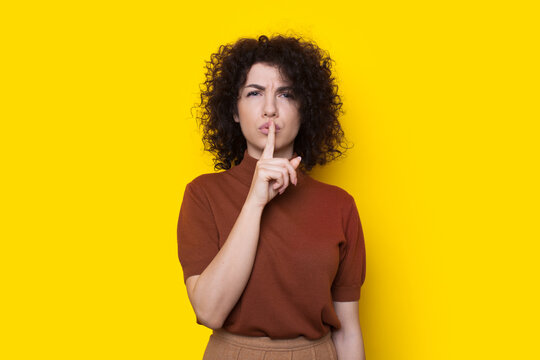 Portrait of serious woman showing silence gesture with finger over lips isolated over yellow background. Shh sign. Curly hair.