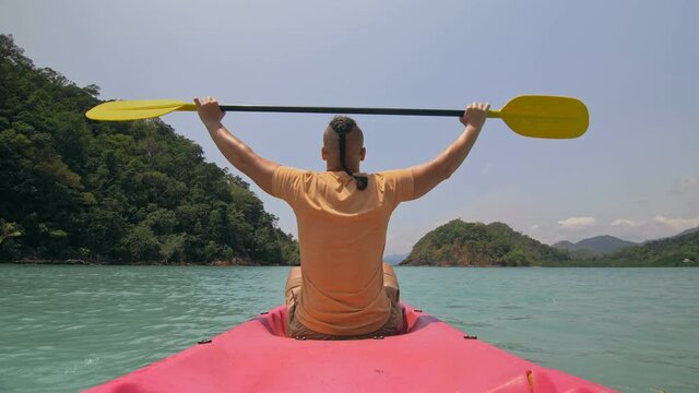 Man with thin plait hairstyle rows pink plastic kayak putting up paddle on sea against hills with wild jungles and blue sky.