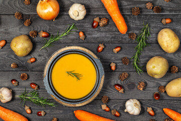 Carrot and potato soup on a black wood table with vegetables, acorns, pine cones, garlic and rosemary. Flat lay.