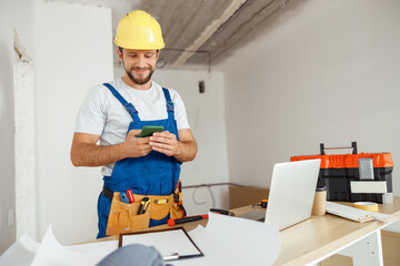 Joyful handyman in workwear and hardhat smiling while using smartphone, standing indoors during...