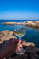 Woman's hand holding glass of white dry white wine with view on rocks and blue sea bay water near Protaras touristic town on Cyprus