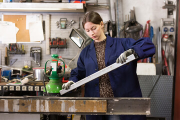 Confident young woman in blue overalls working in mechanical metalworking workshop, checking metal...