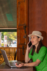Latin woman with hat and green blouse sitting in a coffee shop working on her laptop while talking on her cell phone