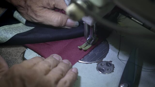 Close-up of hands of an elderly tailor who is sewing a brown lining to a leather jacket on sewing machine in a beam of light in a dark workshop. A master with a beautiful big ring with a purple stone