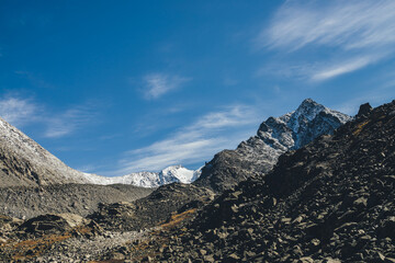 Awesome alpine landscape with dark rockies and high snow-covered mountains in sunshine under blue...