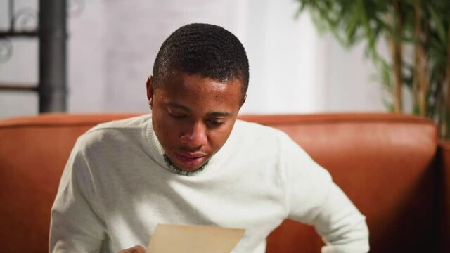 Confused African-American guy in white polo neck sweater looks at old picture of family members with regret sitting on couch at home closeup slow motion