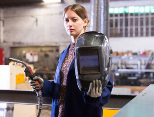 Portrait of young female metalworker standing with welding machine and protective helmet in...