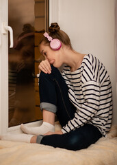 Obraz premium A cute teenage girl sits on a window listening to music with headphones and thinking.