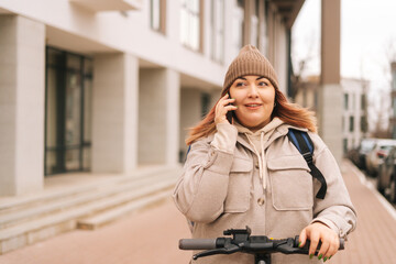 Female courier food delivery with big thermo backpack on back talking on phone with client standing with electric scooter in urban street. Delivery woman in warm outerwear delivering order to customer