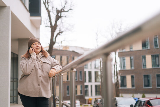 Focused Pretty Overweight Woman In Warm Hat And Jacket Talking On Mobile Phone Standing Near Railing At City Street In Cloudy Autumn Day. Serious Obese Female Having Call On Smartphone Outdoors.