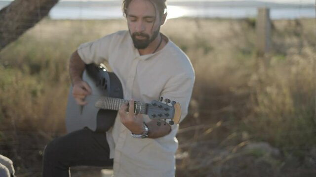 Hispanic bearded hipster man playing  guitar by the road in countryside - pull back, slow-motion