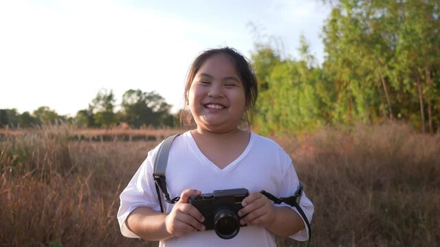 Front view of happy Aisan girl standing in meadow and holding a camera, smiling and looking at camera. Standing on grass in beautiful day with blue sky background