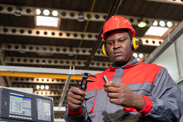 CNC Machine Operator In Protective Workwear Checking Measurements With Vernier Caliper. Portrait Of Black Worker In Red Helmet, Hearing Protectors And Work Uniform Next To CNC Controller.