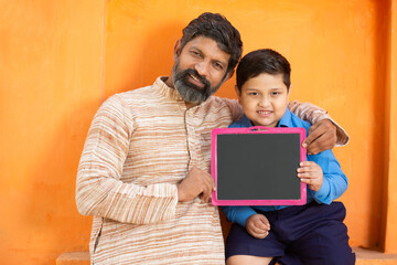 Happy indian father and his cute little son in school uniform holding blank slate against orange...