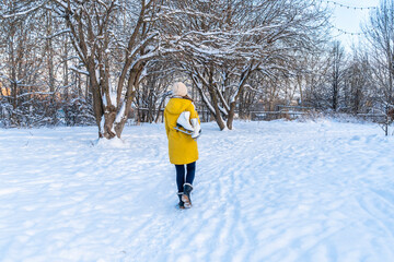 Young woman in yellow jacket with ice skates walking through snowy park. View from behind.