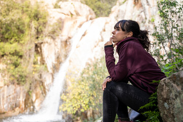Pensive woman sitting while looking at a small waterfall in the forest