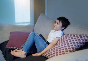 Happy boy with smiling face sitting on sofa watching cartoon, School Kid sitting alone on couch holding tablet and watching TV, Portrait Child relaxing in living room after back from school.