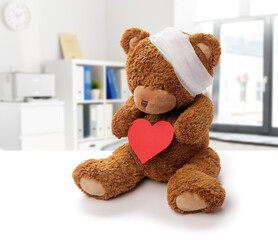 medicine, healthcare and childhood concept - teddy bear toy with bandaged head holding red heart over hospital background