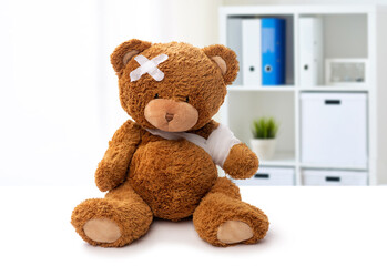 medicine, healthcare and childhood concept - teddy bear toy with bandaged paw and patch on head over medical office at hospital background