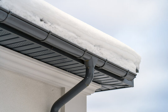 Corner of house with new roof made of gray metal tiles and gutter covered with thick layer of snow in winter. Metal Downpipe system, Guttering System, External downpipes and drainage pipes