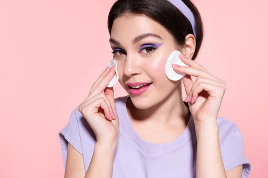 Beautiful modern young girl cleansing face, using cotton pads. Stylish female removing trendy lavender makeup by cotton pad on pink studio background. Skincare daily procedure.