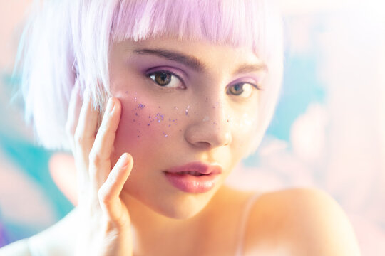 Young modern blond woman with perfect hydrated skin and sparkles on face. Beautiful sensual blonde girl with short bob haircut touch cheek applying glitters. Skincare and contemporary makeup.