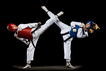 Dynamic portrait of two young women, taekwondo athletes training together isolated over dark...