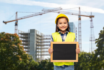 building and profession concept - smiling little girl in protective helmet and safety vest holding chalkboard over construction site background