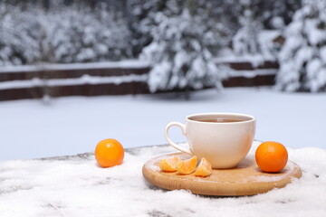 A white mug of hot tea is among tangerines on a snowy wooden table on the winter forest background.
