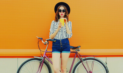 Summer colorful portrait of young woman drinking a juice with bicycle in the city on orange...