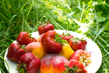 Strawberries and peaches on a plate on the green grass. Delicious and fresh summer fruits. Healthy vegetarian food with vitamins.