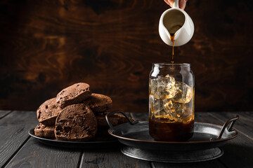 Making iced coffee. Pouring black coffee from a milk jug into a glass. Chocolate cookies on a plate.