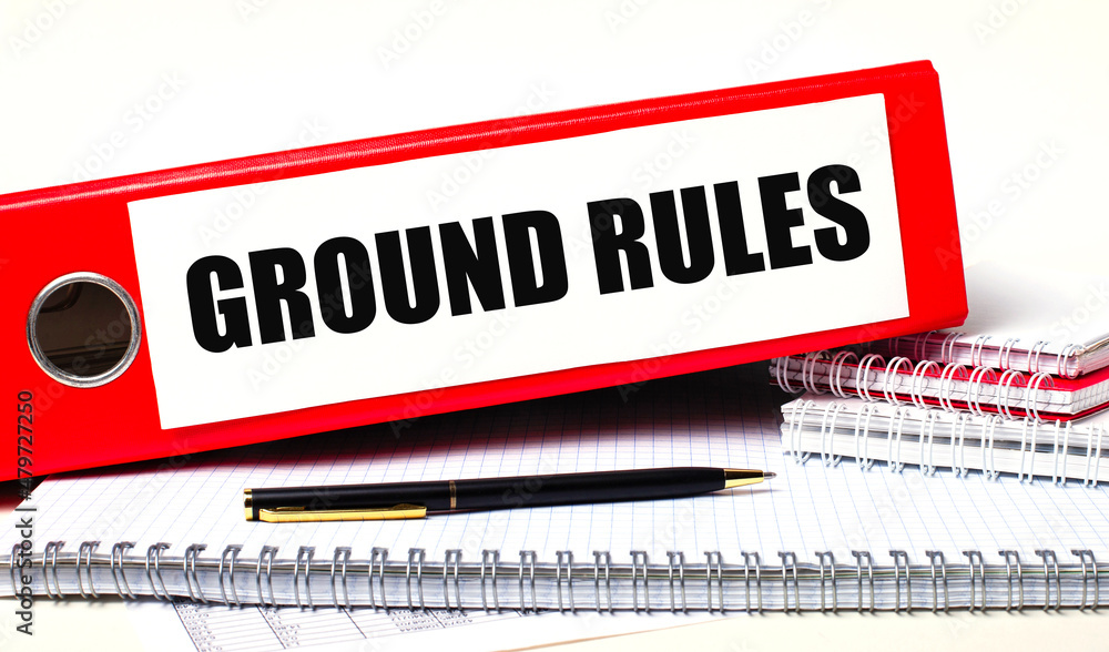 Poster on the desktop are notebooks, a pen and a red folder for papers with the text ground rules. business