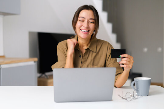 Excited Asian woman shopping online sitting with credit card at home office, looking at the laptop screen and rejoicing to purchase, multiracial female making order online, booking