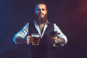 Hipster man holds dumbbell and mug of beer in hands. Concept of choice between alcohol and sport