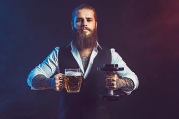 Fototapeten Hipster man holds dumbbell and mug of beer in hands. Concept of choice between alcohol and sport © zamuruev