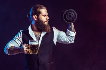 bearded man holds dumbbell and mug of beer on dark background while choices alcohol or sport
