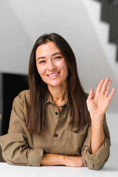 Young smiling Asian female on an online interview or making video call, sitting at desk and looking waving at camera, saying hello, woman takes a part in virtiual meeting. Job hunting. Vertical shot
