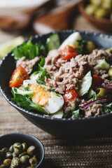 Canned tuna salad with fresh vegetables, capers and olives in a black bowl. Healthy lunch or dinner.