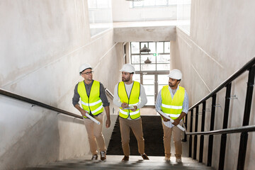 architecture, construction business and people concept - male architects in helmets with blueprints and clipboard walking upstairs at office building