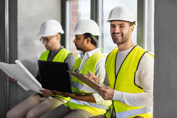architecture, construction business and people concept - group of male architects in helmets with laptop, blueprint and clipboard working at office