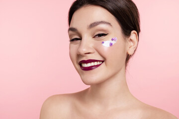 Pretty young woman with perfect make up, smooth healthy skin, cream with flowers on cheek and bare...