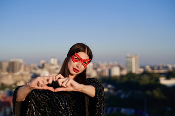 Make love, not war. Portrait of young brunette superwoman in black dress and red face mask making heart shape with hands on urban background. Freedom and equality, female power, women rights concept