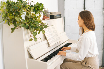 Talented attractive young woman musician performer playing gentle music on white classical piano in light home studio. Female student pianist practicing in classroom using musical notes. .