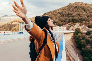 Young woman arms outstretched by the mountain at sunrise enjoying freedom and life, people travel wellbeing concept. Cheerful smiling woman wearing winter clothes enjoying at the top of dam.
