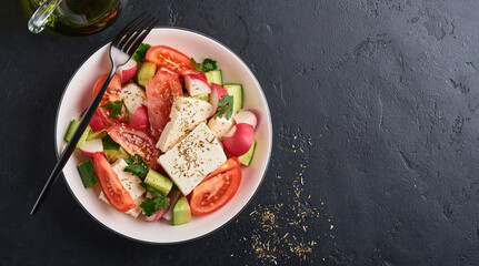Radish, cucumber, tomato, pepper and feta cheese with spices pepper and olive oil in white bowl on black slate, stone or concrete background. Healthy food concept. Top view.