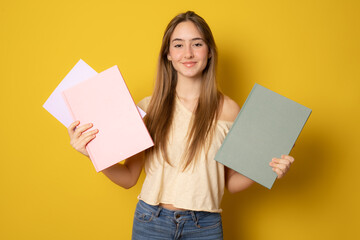 Portrait of a cute young student girl holding colorful notebooks isolated on yellow background - obrazy, fototapety, plakaty